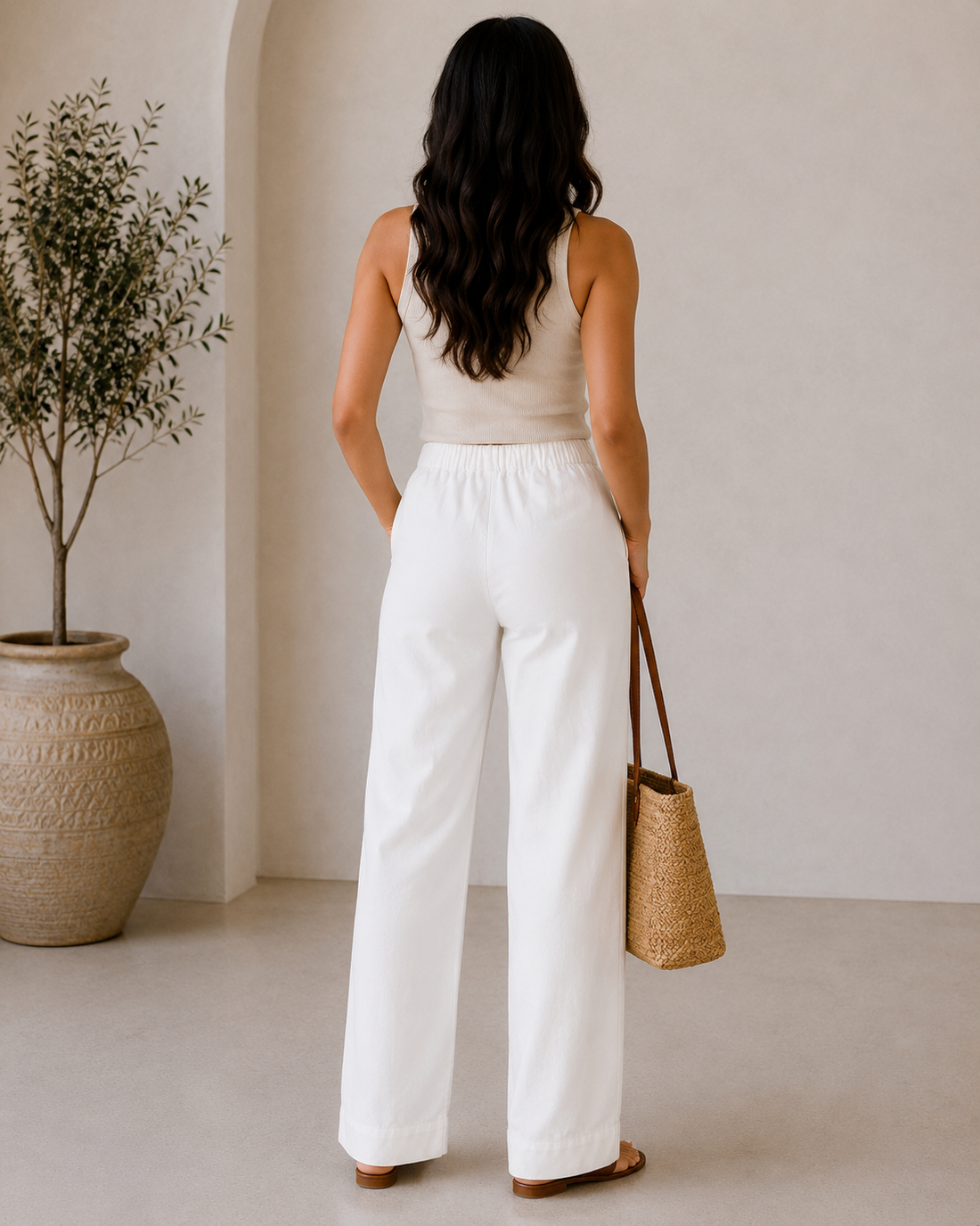 Woman in a beige top and white pants holding a straw bag in a minimalistic room.