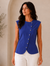 A woman modeling a sleeveless royal blue vest with decorative buttons down the front, paired with white wide-leg pants and brown sandals, in a warm, minimalist interior setting.