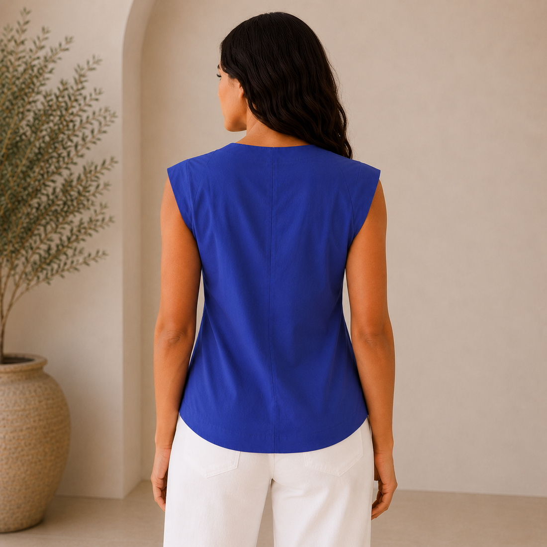 A woman modeling a sleeveless royal blue vest with decorative buttons down the front, paired with white wide-leg pants and brown sandals, in a warm, minimalist interior setting.