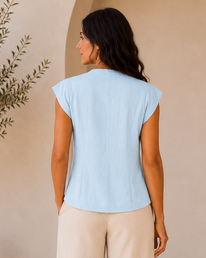 Woman wearing a light blue top and beige pants standing in a room with a plant and arches.