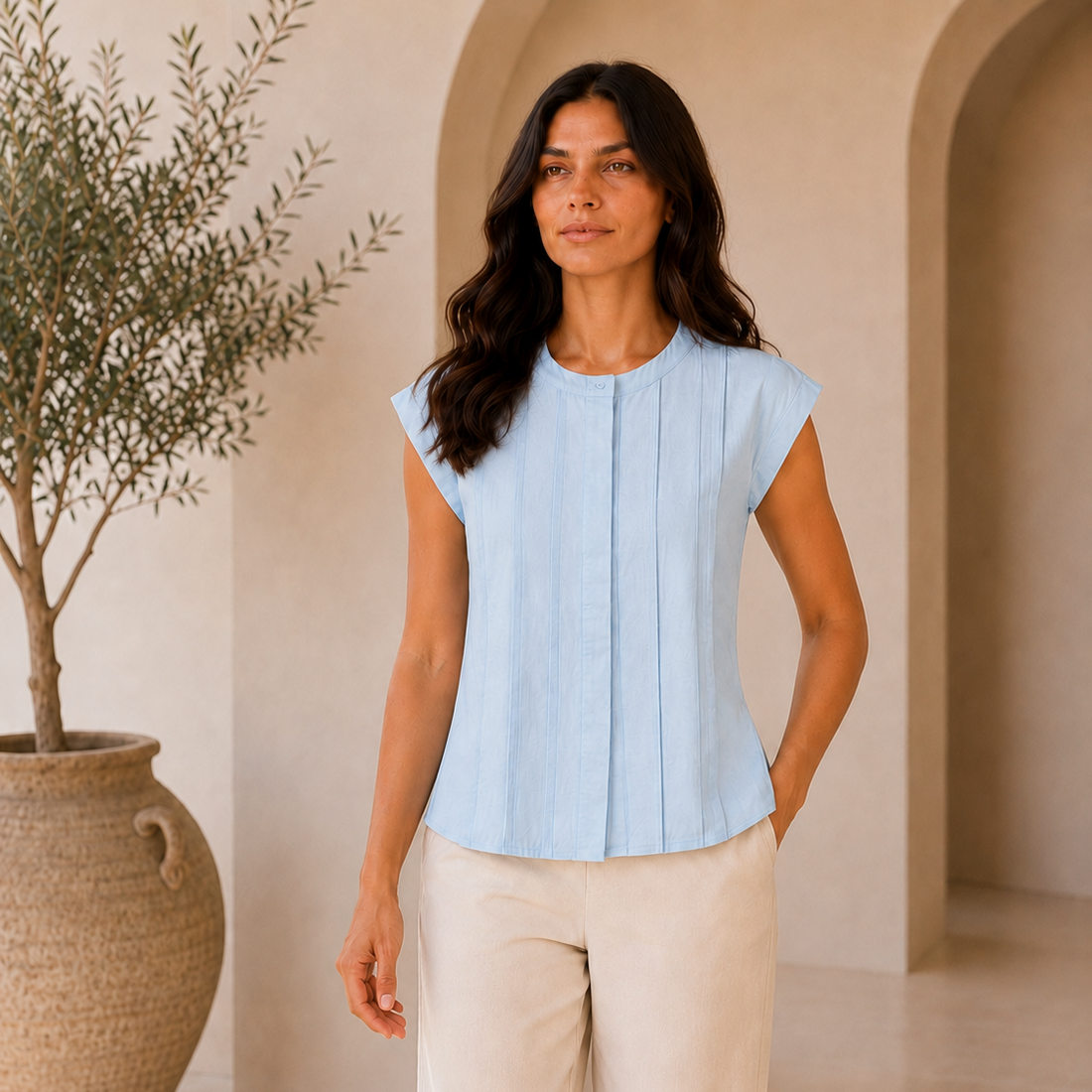 Woman wearing a light blue top and beige pants standing in a sunlit room with a plant.