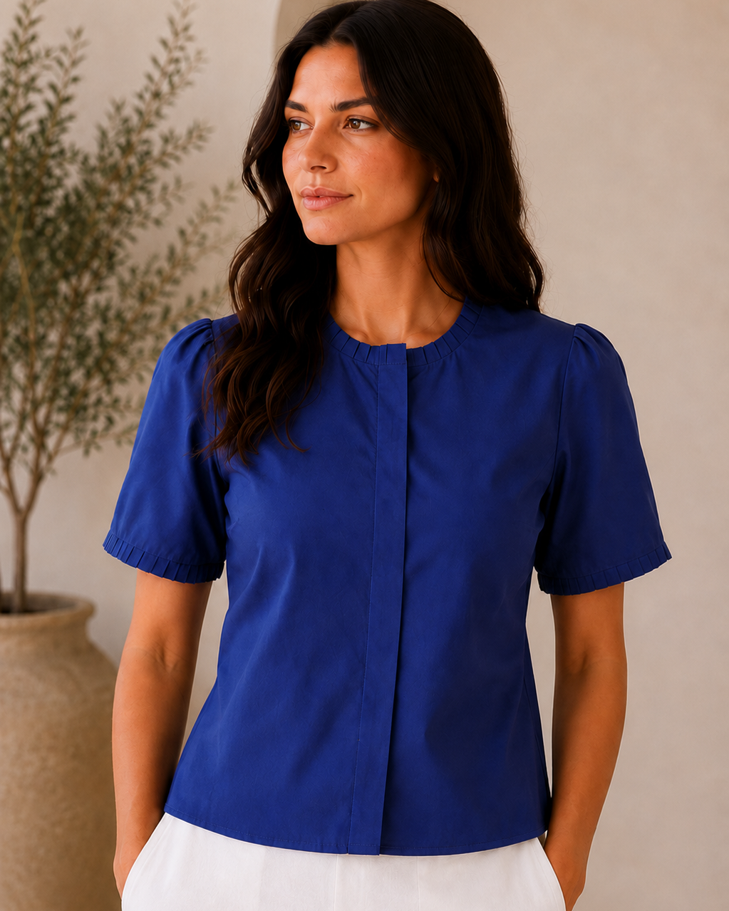 A woman wearing a royal blue short-sleeve blouse with a round neckline and front button placket, paired with white pants, standing in a softly lit indoor setting.