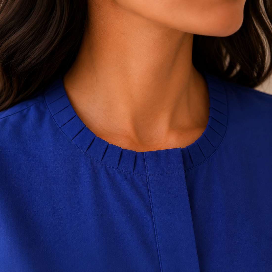 A woman wearing a royal blue short-sleeve blouse with a round neckline and front button placket, paired with white pants, standing in a softly lit indoor setting.